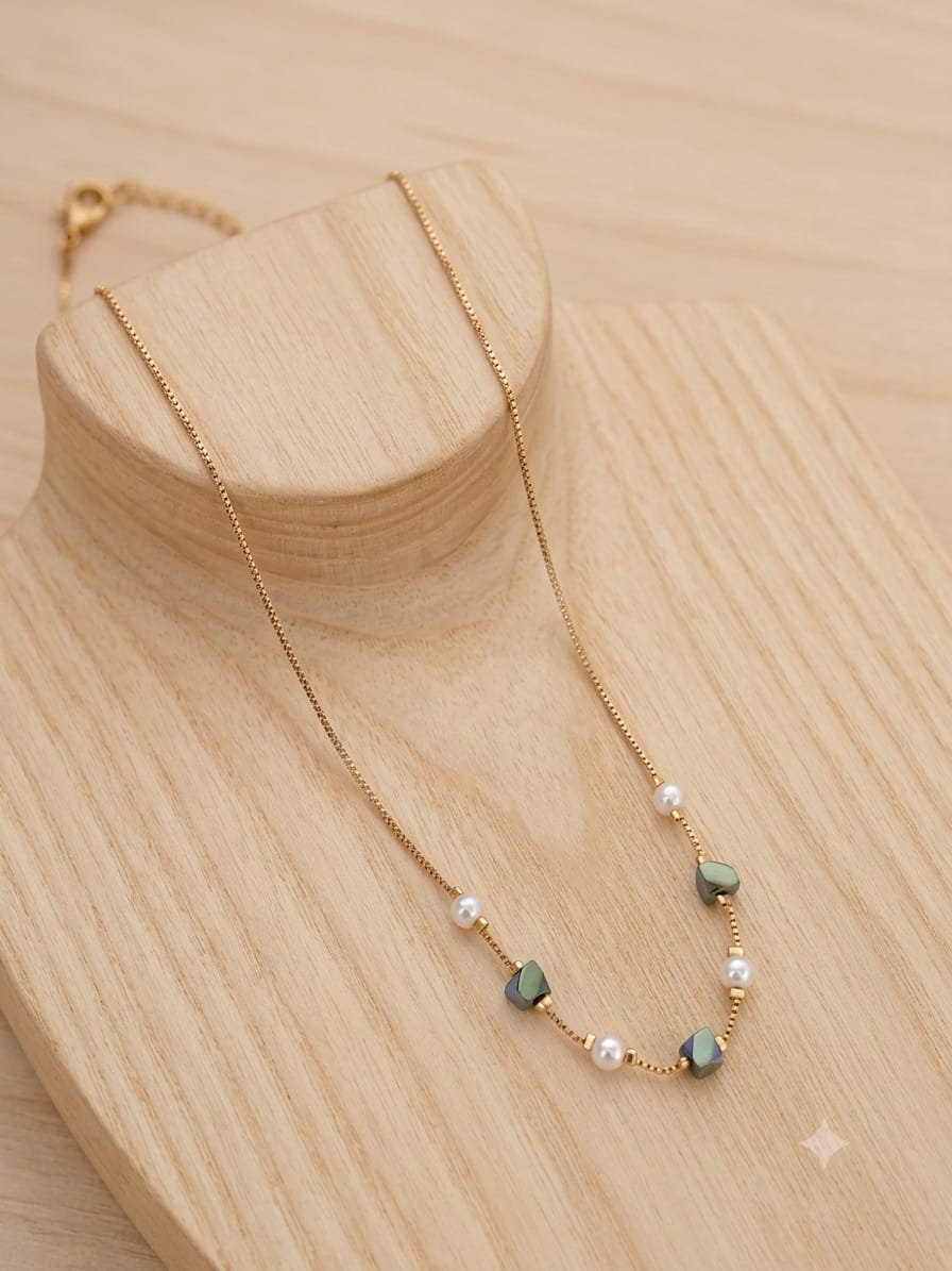 A delicate gold chain necklace with small white pearls and green faceted beads, displayed on a light wood stand against a matching wood background.