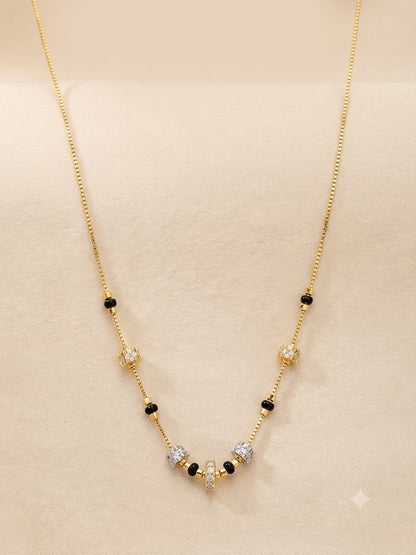 A delicate gold chain necklace with small black beads and a central cluster of clear and gold-toned stones, displayed on a soft beige jewelry stand.