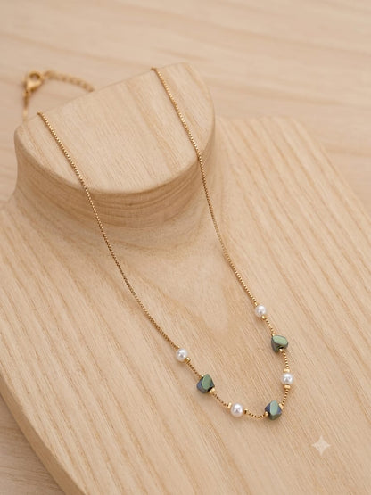 A delicate gold chain necklace with small white pearls and green faceted beads, displayed on a light wood stand against a matching wood background.